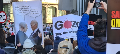 A crowd of protesters holding various signs, including a satirical poster depicting a conversation between two political figures with speech bubbles about bombing and financial support. Other signs include messages and slogans about the situation in Gaza. The scene suggests a public demonstration with participants capturing images.