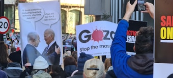 A crowd of protesters holding various signs, including a satirical poster depicting a conversation between two political figures with speech bubbles about bombing and financial support. Other signs include messages and slogans about the situation in Gaza. The scene suggests a public demonstration with participants capturing images.