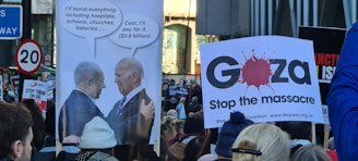 A protest scene with people holding signs. One sign features two political leaders with speech bubbles discussing bombing, while another indicates a plea to 'Stop the massacre' in Gaza. The crowd appears engaged, with various messages visible on placards.