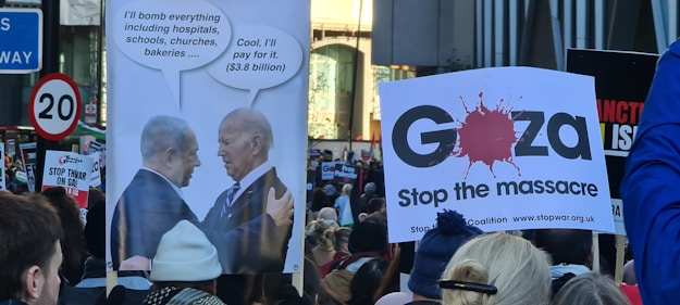 A protest scene with people holding signs. One sign features two political leaders with speech bubbles discussing bombing, while another indicates a plea to 'Stop the massacre' in Gaza. The crowd appears engaged, with various messages visible on placards.