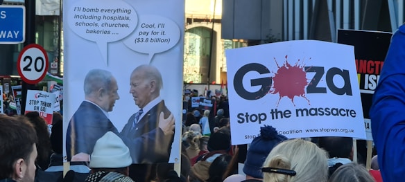 A protest scene with people holding signs. One sign features two political leaders with speech bubbles discussing bombing, while another indicates a plea to 'Stop the massacre' in Gaza. The crowd appears engaged, with various messages visible on placards.
