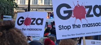 Crowd at a protest holding signs with messages advocating to stop the massacre in Gaza, featuring a large red splatter graphic. Policemen and buildings are visible in the background.