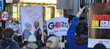 A crowd of people is participating in a protest, holding various signs and placards. Some signs contain slogans advocating for political change, while others display satirical cartoons. The scene is lively, with a mix of banners and diverse individuals.