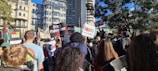A large crowd gathers in an urban setting, with people holding signs and banners advocating for political causes. The backdrop includes modern and historical buildings under a clear blue sky. Trees are visible, adding a touch of nature to the city environment.