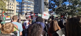 A large crowd gathers in an urban setting, with people holding signs and banners advocating for political causes. The backdrop includes modern and historical buildings under a clear blue sky. Trees are visible, adding a touch of nature to the city environment.