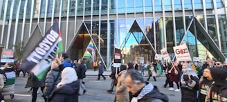 A group of people are marching on the street, holding various signs and flags. The signs carry messages about boycotting and supporting Palestine. The setting appears to be an urban environment with a large modern glass building in the background.