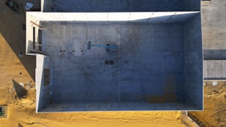 An aerial view of a large, rectangular, partially constructed building. The interior is empty, with a small vehicle, likely a construction crane or lift, positioned in the center. The building is surrounded by sandy terrain, and shadows suggest late afternoon lighting.