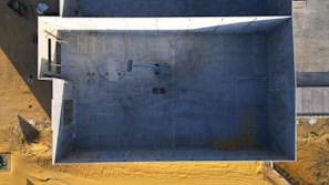 An aerial view of a large, rectangular, partially constructed building. The interior is empty, with a small vehicle, likely a construction crane or lift, positioned in the center. The building is surrounded by sandy terrain, and shadows suggest late afternoon lighting.