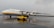 A large Antonov cargo aircraft is parked on a wet runway at an airport. It is white with blue and yellow accents and has four large jet engines. A yellow airport service vehicle is positioned in front of the plane.