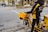 A person wearing a black and yellow jacket stands beside a yellow delivery bicycle on a city street. The bicycle is equipped with a large yellow box and carrying bags, indicating it's used for postal or delivery services. Nearby, a few cars and pedestrians are visible, along with trees lining the street.