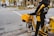 A person wearing a black and yellow jacket stands beside a yellow delivery bicycle on a city street. The bicycle is equipped with a large yellow box and carrying bags, indicating it's used for postal or delivery services. Nearby, a few cars and pedestrians are visible, along with trees lining the street.