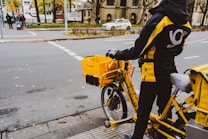 A person wearing a black and yellow jacket stands beside a yellow delivery bicycle on a city street. The bicycle is equipped with a large yellow box and carrying bags, indicating it's used for postal or delivery services. Nearby, a few cars and pedestrians are visible, along with trees lining the street.