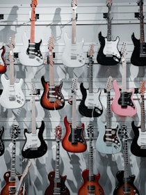A display of electric guitars hanging on a wall, featuring a variety of designs and colors including white, black, pink, red, and sunburst finishes. The guitars are arranged in rows on slat wall mounts, showcasing different body styles and headstocks.