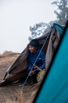 A person sits partially inside a makeshift tarp shelter on a barren, grassy landscape. The individual wears a blue jacket and leans forward with hands clasped. A woven basket sits next to them. In the background, blurred foliage is visible, suggesting an outdoor, possibly mountainous environment.