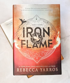 A hardcover book titled 'Iron Flame' by Rebecca Yarros, featuring an ornate design with a prominent black and white title against a fiery orange and red background. An illustration of a flying dragon is present, set within intricate cloud and flame patterns. The top of the cover includes a quote praising the book, attributed to #1 NYT bestselling author Jennifer L. Armentrout.