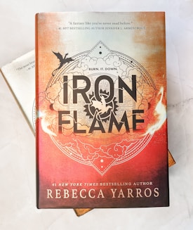 A hardcover book titled 'Iron Flame' by Rebecca Yarros, featuring an ornate design with a prominent black and white title against a fiery orange and red background. An illustration of a flying dragon is present, set within intricate cloud and flame patterns. The top of the cover includes a quote praising the book, attributed to #1 NYT bestselling author Jennifer L. Armentrout.