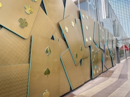 A wall decorated with large metallic playing cards, each featuring various suit symbols such as clubs, spades, diamonds, and hearts. The cards are arranged in a staggered, overlapping pattern against a backdrop of modern glass architecture.