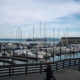 The image depicts a marina filled with numerous sailboats and yachts docked in the water. In the background, a large container ship is seen passing by on the water, and there is a mountain range in the distance. The sky is cloudy with some blue patches. In the foreground, a person wearing a cap is pushing a stroller along a wooden boardwalk.
