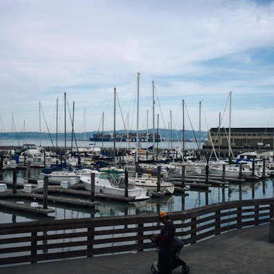The image depicts a marina filled with numerous sailboats and yachts docked in the water. In the background, a large container ship is seen passing by on the water, and there is a mountain range in the distance. The sky is cloudy with some blue patches. In the foreground, a person wearing a cap is pushing a stroller along a wooden boardwalk.