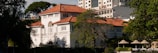 A large, elegant two-story colonial-style house with a red-tiled roof and white walls. The house is surrounded by trees and vegetation, with modern high-rise buildings visible in the background. Several parasols and lounge chairs are arranged neatly in an outdoor area in front of the house.