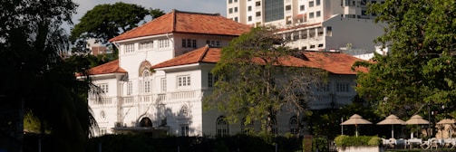 A large, elegant two-story colonial-style house with a red-tiled roof and white walls. The house is surrounded by trees and vegetation, with modern high-rise buildings visible in the background. Several parasols and lounge chairs are arranged neatly in an outdoor area in front of the house.