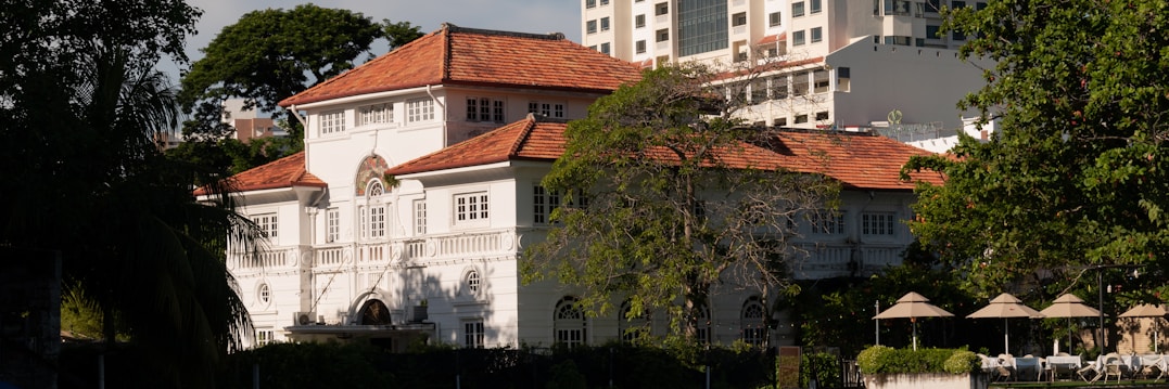 A large, elegant two-story colonial-style house with a red-tiled roof and white walls. The house is surrounded by trees and vegetation, with modern high-rise buildings visible in the background. Several parasols and lounge chairs are arranged neatly in an outdoor area in front of the house.
