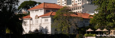 A large, elegant two-story colonial-style house with a red-tiled roof and white walls. The house is surrounded by trees and vegetation, with modern high-rise buildings visible in the background. Several parasols and lounge chairs are arranged neatly in an outdoor area in front of the house.