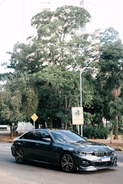 A sleek, dark-colored sedan is parked on a city street. Behind it, lush green trees provide a natural backdrop, and urban buildings peek through the foliage. Road signs, including a pedestrian crossing warning with text in Portuguese, are visible.