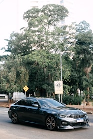 A sleek, dark-colored sedan is parked on a city street. Behind it, lush green trees provide a natural backdrop, and urban buildings peek through the foliage. Road signs, including a pedestrian crossing warning with text in Portuguese, are visible.