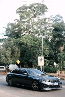 A sleek, dark-colored sedan is parked on a city street. Behind it, lush green trees provide a natural backdrop, and urban buildings peek through the foliage. Road signs, including a pedestrian crossing warning with text in Portuguese, are visible.