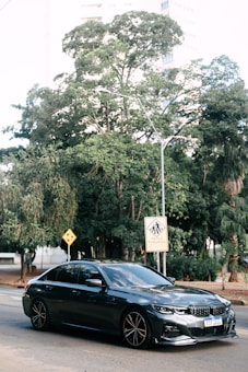 A sleek, dark-colored sedan is parked on a city street. Behind it, lush green trees provide a natural backdrop, and urban buildings peek through the foliage. Road signs, including a pedestrian crossing warning with text in Portuguese, are visible.