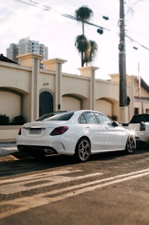 A white luxury sedan is parked on a street in front of a beige building with arched windows and palm trees in the background. The lighting suggests late afternoon, with a clear sky and modern city buildings visible in the distance.