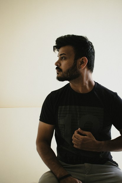 A person is seated against a plain light-colored background, wearing a dark t-shirt with a subtle graphic design. The person has short, styled hair and a beard, looking pensively to the side. Their posture is relaxed, with one hand resting on their chest.