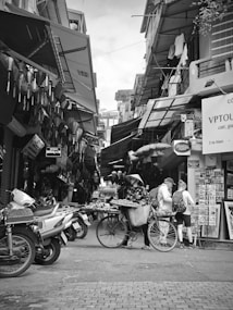 A bustling narrow street scene in a market with various stalls and signs. A person wearing a conical hat is pushing a bicycle laden with goods for sale. Two people with backpacks are looking at a display outside a shop. Several motorbikes are parked along the street. The atmosphere suggests a typical market day in an urban area.