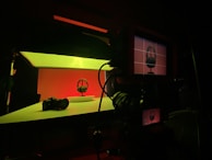 A photography setup with a pair of headphones on a small pedestal, illuminated with a vibrant red and green lighting scheme. In the foreground, a camera is positioned to capture the scene, and a monitor displays a preview of the shot. The surrounding area is dimly lit, highlighting the colorful and dramatic lighting of the subject.