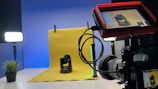 A professional photography setup is visible with a camera mounted on a tripod, pointing towards a yellow backdrop with a small robotic camera placed in front. A monitor screen attached to the camera displays the feed of the robotic camera. Studio lighting equipment is present on both sides, creating a well-lit environment. A small plant is placed to the left on a table.