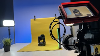A professional photography setup is visible with a camera mounted on a tripod, pointing towards a yellow backdrop with a small robotic camera placed in front. A monitor screen attached to the camera displays the feed of the robotic camera. Studio lighting equipment is present on both sides, creating a well-lit environment. A small plant is placed to the left on a table.