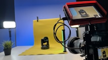 A professional photography setup is visible with a camera mounted on a tripod, pointing towards a yellow backdrop with a small robotic camera placed in front. A monitor screen attached to the camera displays the feed of the robotic camera. Studio lighting equipment is present on both sides, creating a well-lit environment. A small plant is placed to the left on a table.