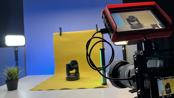 A professional photography setup is visible with a camera mounted on a tripod, pointing towards a yellow backdrop with a small robotic camera placed in front. A monitor screen attached to the camera displays the feed of the robotic camera. Studio lighting equipment is present on both sides, creating a well-lit environment. A small plant is placed to the left on a table.