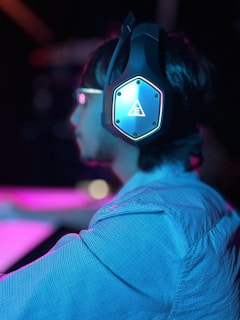 A person is wearing large, illuminated headphones, focusing on something outside the frame. The ambient lighting is a mix of pink and blue hues, creating a vibrant and modern atmosphere.