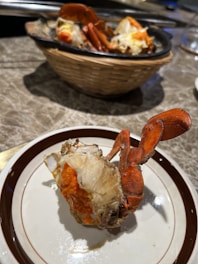 A cooked crab dish is presented on a white plate with brown trim. The vibrant orange crab is served with its shell partially removed, revealing the internal meat and roe. In the background, there is a black dish with more crab pieces resting on a wicker basket, all placed on a marble table.