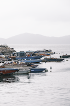 Boats are moored along a calm waterfront, with a rocky shoreline to the left. The background features hazy, rolling hills under an overcast sky. Several boats of varying sizes and colors, including blue and brown ones, are docked in the harbor area.
