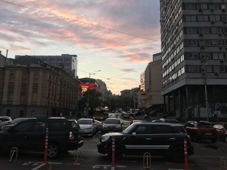 A bustling city street at dusk, framed by tall buildings on either side. The sky is painted with pink and orange hues, suggesting sunset. Several cars are parked along the street, with others driving by. A large advertisement on one of the buildings glows brightly, and a bus can be seen in the distance. Light poles and street signs are visible, adding to the urban atmosphere.