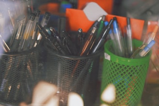 Multiple pens of various colors are placed in two distinct holders, one black and one green, on a desk. The pens include both gel and ballpoint types, with some featuring blue caps. The background contains blurred orange and blue objects, creating a casual and slightly disorganized workspace ambiance.