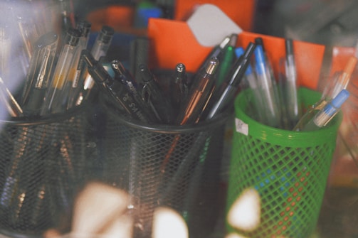 Multiple pens of various colors are placed in two distinct holders, one black and one green, on a desk. The pens include both gel and ballpoint types, with some featuring blue caps. The background contains blurred orange and blue objects, creating a casual and slightly disorganized workspace ambiance.
