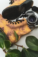 A vintage Nikon camera lies nestled among a pair of rugged, worn-in yellow-brown boots. Black laces contrast with the boots' leather. Fresh green leaves frame the composition, adding a touch of nature to the scene.