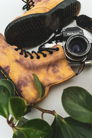 A vintage Nikon camera lies nestled among a pair of rugged, worn-in yellow-brown boots. Black laces contrast with the boots' leather. Fresh green leaves frame the composition, adding a touch of nature to the scene.