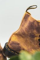 A close-up of a suede boot with a yellow and brown tie-dye pattern is depicted. The boot is detailed with black laces and stitching, highlighting the craftsmanship. The background is slightly blurred, adding focus to the rich texture and color of the shoe.