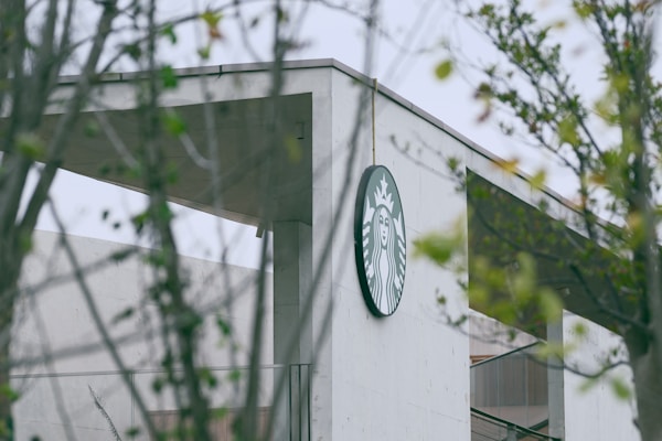 A concrete building is partially obscured by surrounding greenery. The focus is on a round logo featuring a twin-tailed siren, mounted on the upper corner of the building.