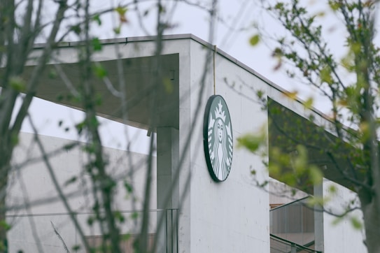 A concrete building is partially obscured by surrounding greenery. The focus is on a round logo featuring a twin-tailed siren, mounted on the upper corner of the building.